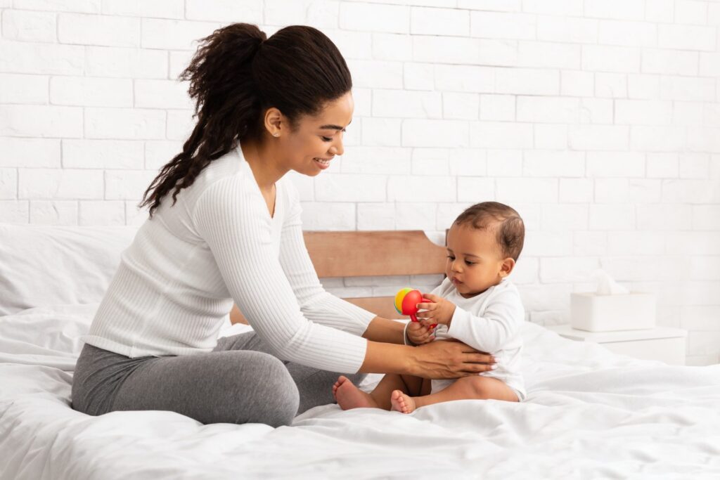 Black mother playing with baby giving toy sitting in bedroom e1634190416345.jpg